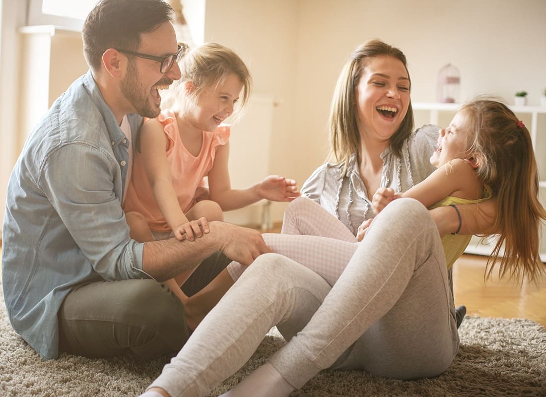 Personal Insurance - Portrait of Cheerful Parents Having Fun Playing with Their Two Daughters at Home While Sitting on a Rug in the Living Room